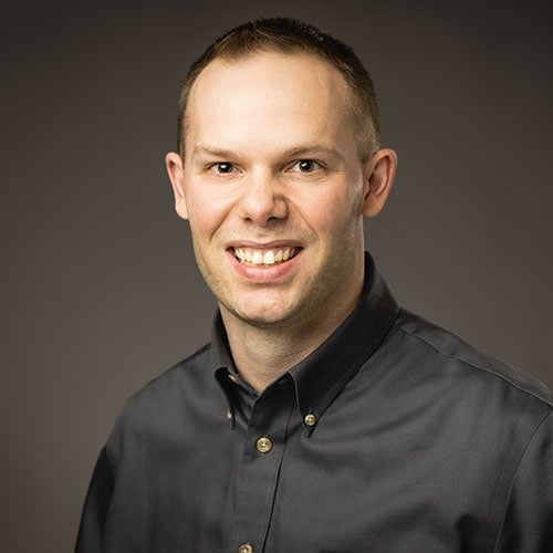 A professional headshot of a smiling man with short hair, wearing a black collared shirt, set against a neutral background, suitable for orthodontic or dental practice websites.