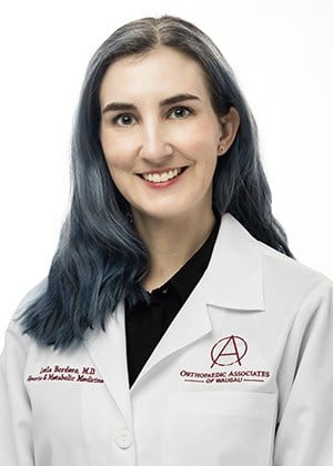 A smiling female orthodontist in a white lab coat, representing professional orthodontic care at Orthodontic Associates of Waukee, with dark blue hair and warm expression.