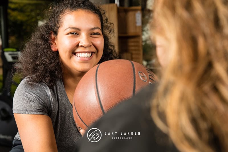 Smiling young woman playing basketball outdoors during the day, holding a basketball with a friendly expression; active lifestyle and sports photography.