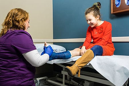 A young girl receiving a pediatric ankle brace fitting at a dentist or medical clinic with a healthcare professional in purple scrubs, sitting on an examination table.