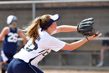 A female softball player in a white jersey and navy cap catches a ball with a glove during a game, with an athlete in the background and a chain-link fence behind her.